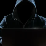 Hacker đội mũ trùm đầu trước máy tính, minh họa nguy cơ lừa đảo (phishing) và tấn công mạng từ trình quản lý mật khẩu.