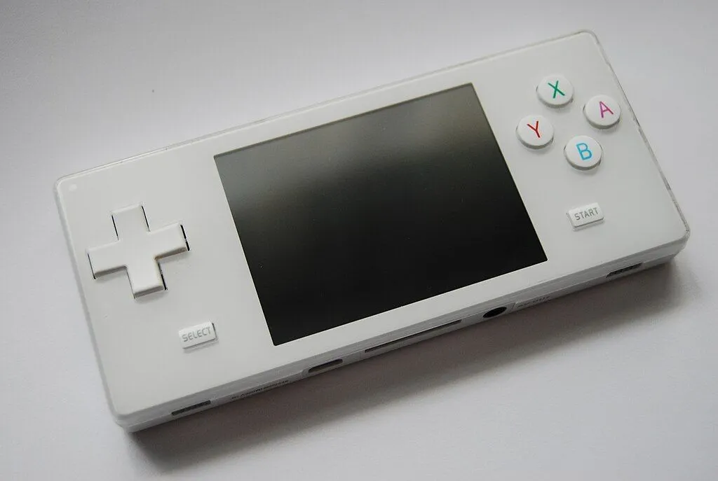 Máy chơi game giả lập Dingoo A320 màu trắng nhỏ gọn với màn hình và các nút điều khiển, đại diện cho dòng console cầm tay đa năng có TV-out.
