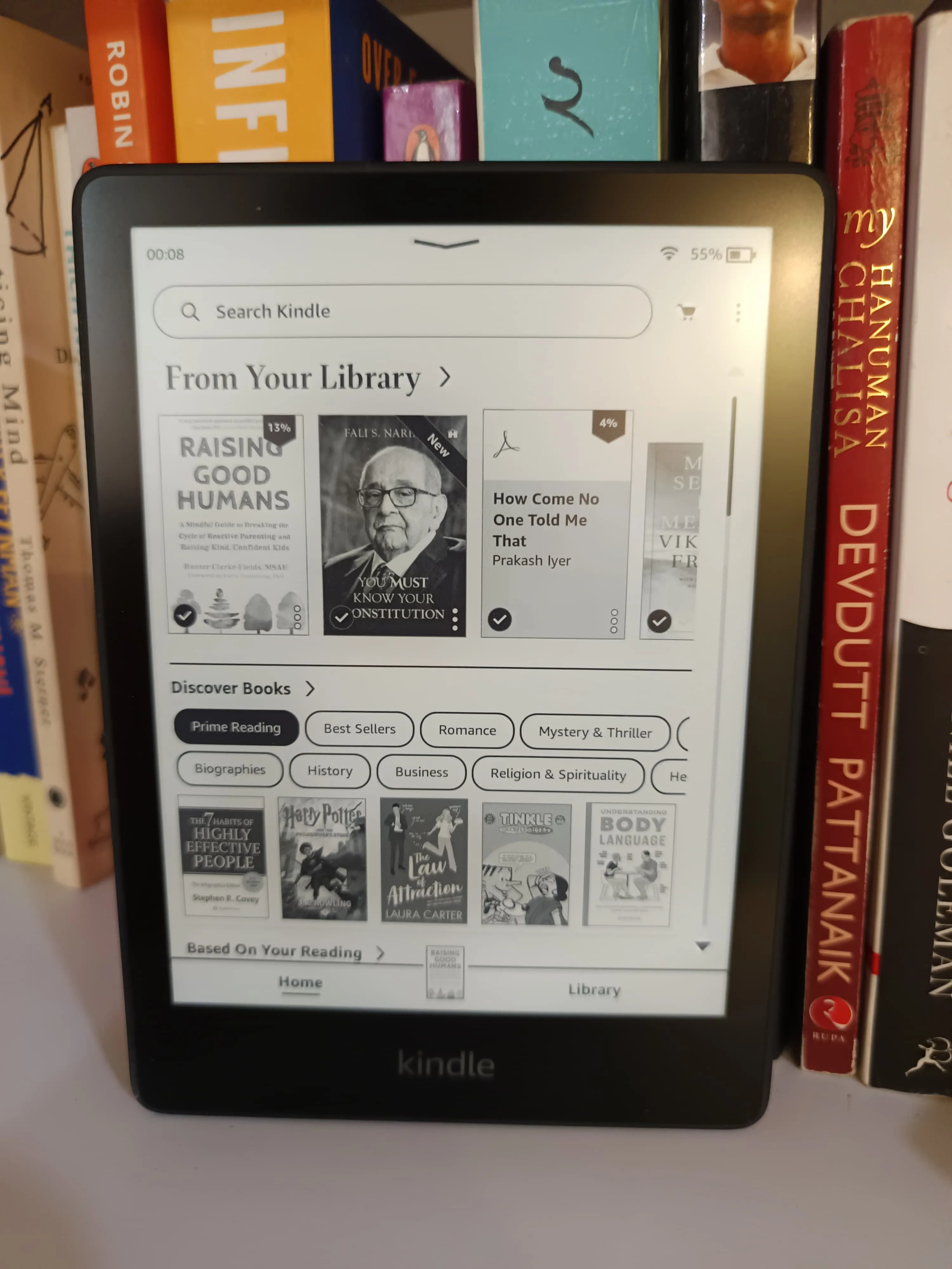 Máy đọc sách Kindle màu đen đặt trên kệ sách gỗ cùng với các cuốn sách, thể hiện vai trò là thiết bị đọc chuyên dụng giúp cải thiện chất lượng giấc ngủ.