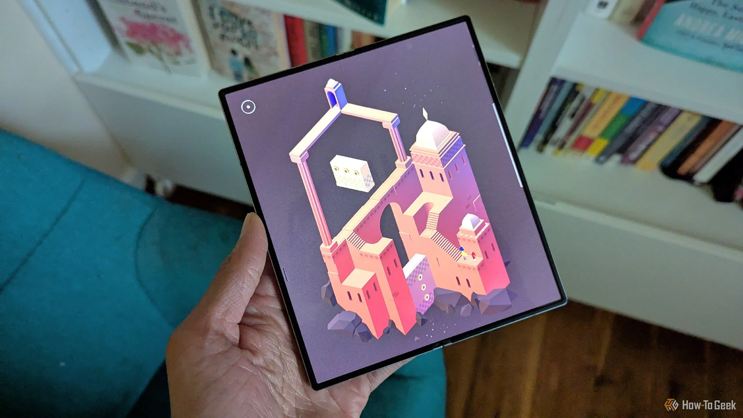 Một chiếc Galaxy Z Fold 6 đang hiển thị trò chơi Monument Valley 2, phía sau là những cuốn sách, tượng trưng cho sự kết hợp giữa công nghệ màn hình gập và trải nghiệm giải trí đa dạng.