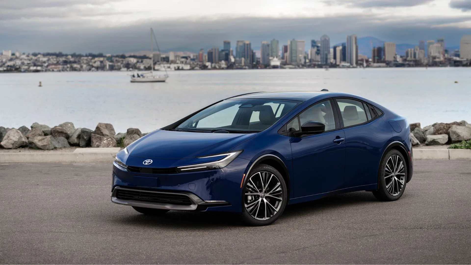 Tổng quan phía trước ba phần tư của một chiếc Toyota Prius 2025 màu xanh hải quân với mặt nước và đường chân trời thành phố phía sau.
