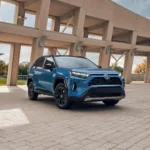 Toyota RAV4 Hybrid 2024 màu xanh lam đỗ trên nền đá cuội với kiến trúc phía sau.