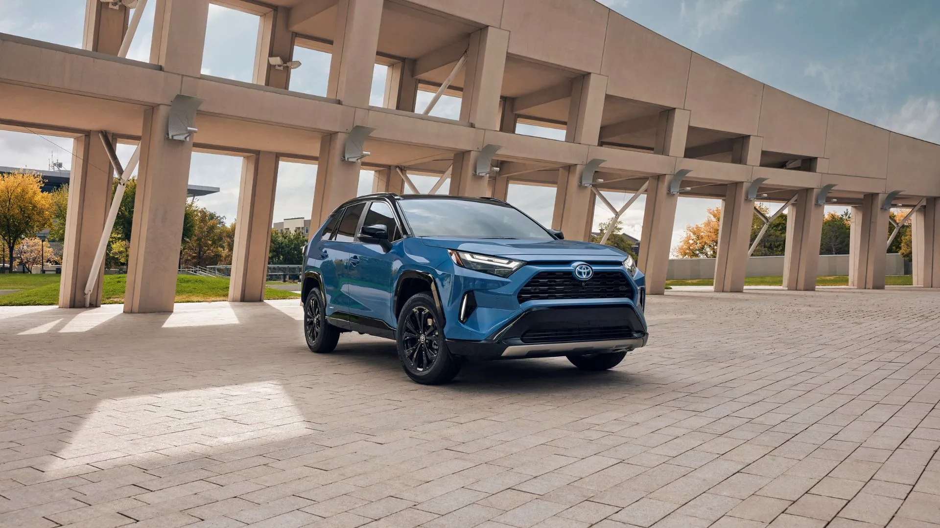 Toyota RAV4 Hybrid 2024 màu xanh lam đỗ trên nền đá cuội với kiến trúc phía sau.