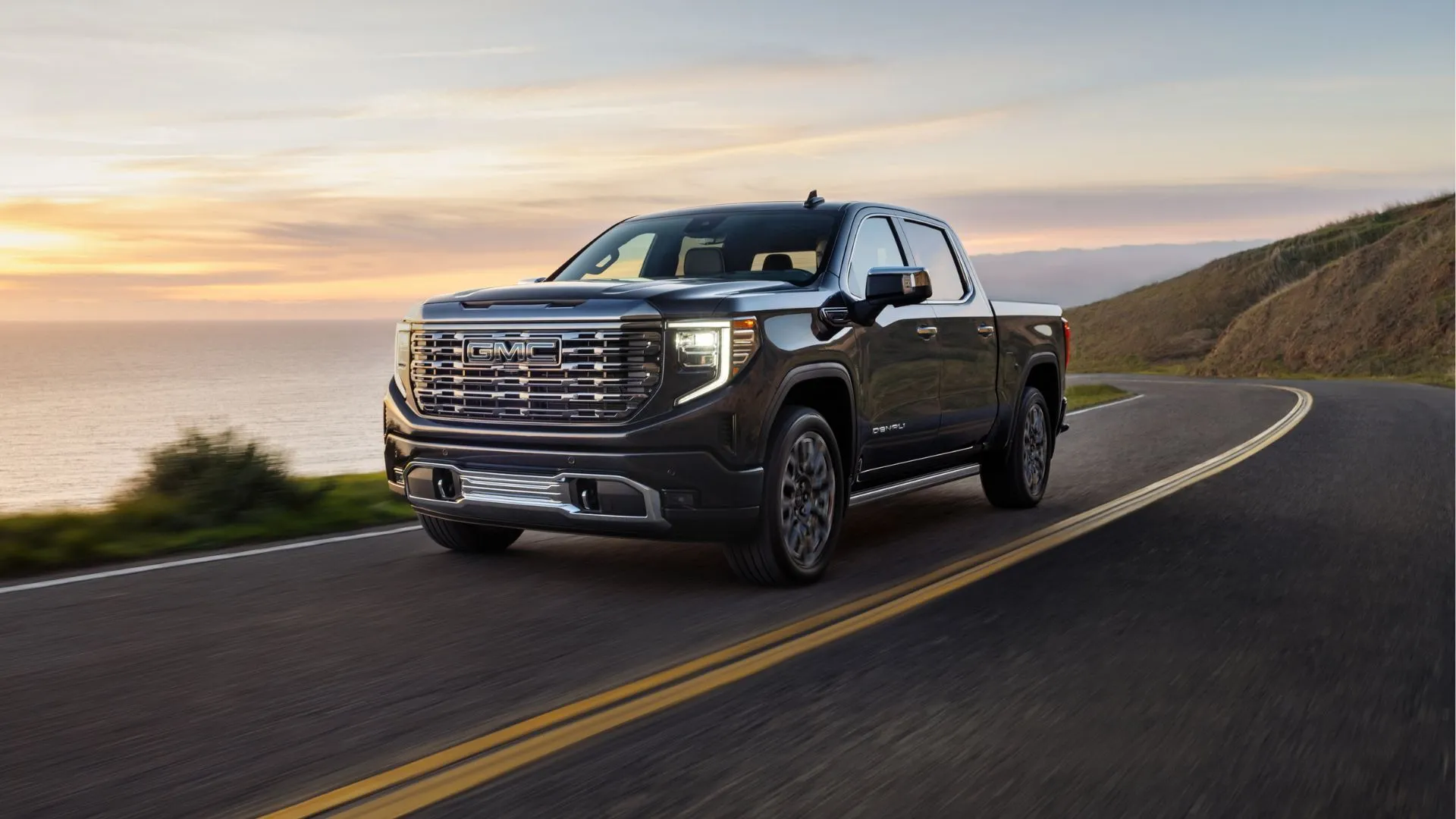 Xe GMC Sierra Denali Ultimate 2025 màu trắng bạc đang di chuyển trên con đường ven biển, biểu tượng cho sức mạnh nhưng cũng tiềm ẩn vấn đề tiêu thụ nhiên liệu do hệ thống DFM bị vô hiệu hóa.